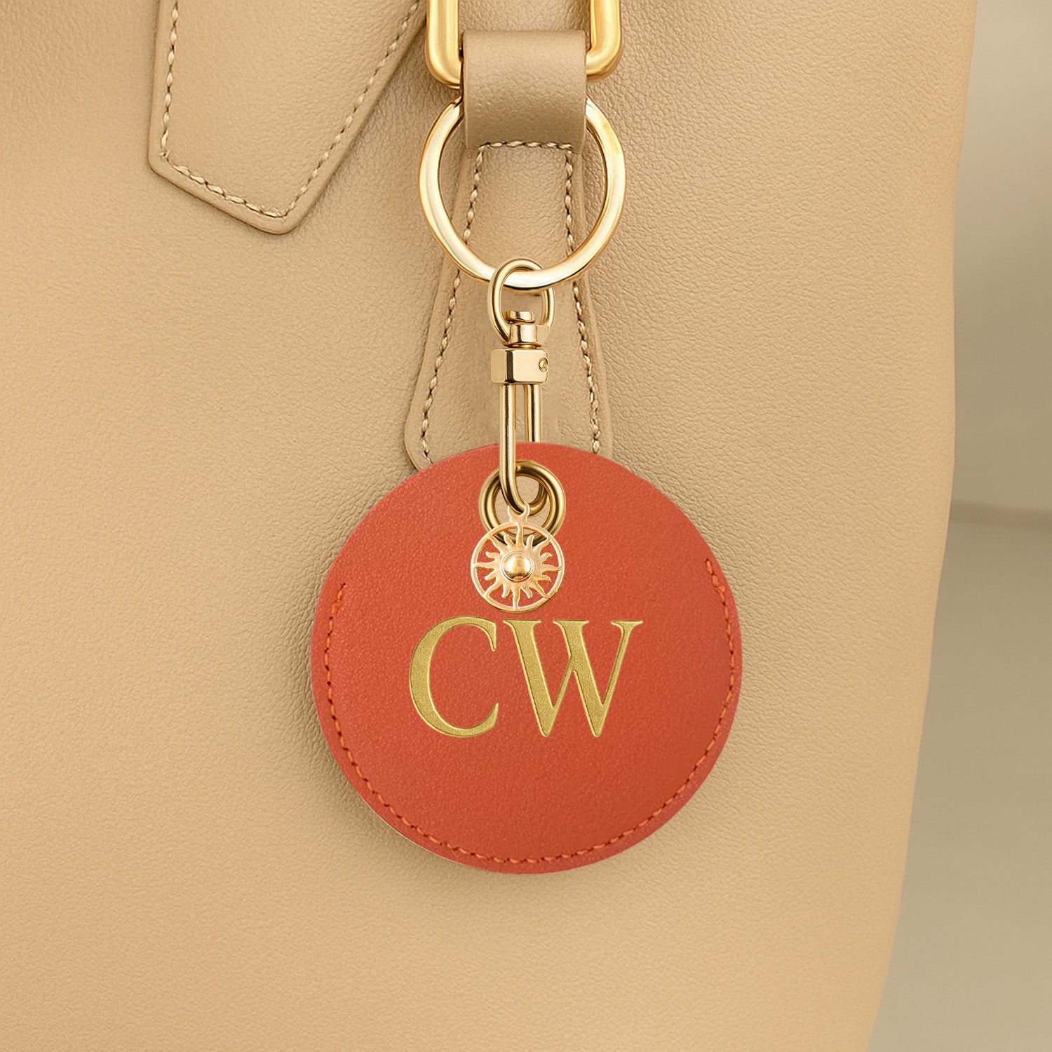 Round red leather keychain with gold initials attached to a beige handbag.
