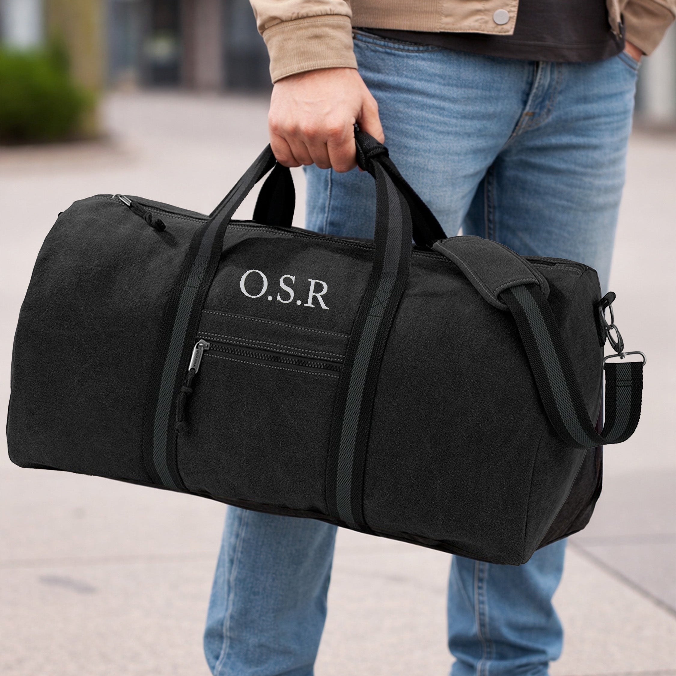 Person holding a black duffel bag with 'O.S.R.' initials on a sidewalk.